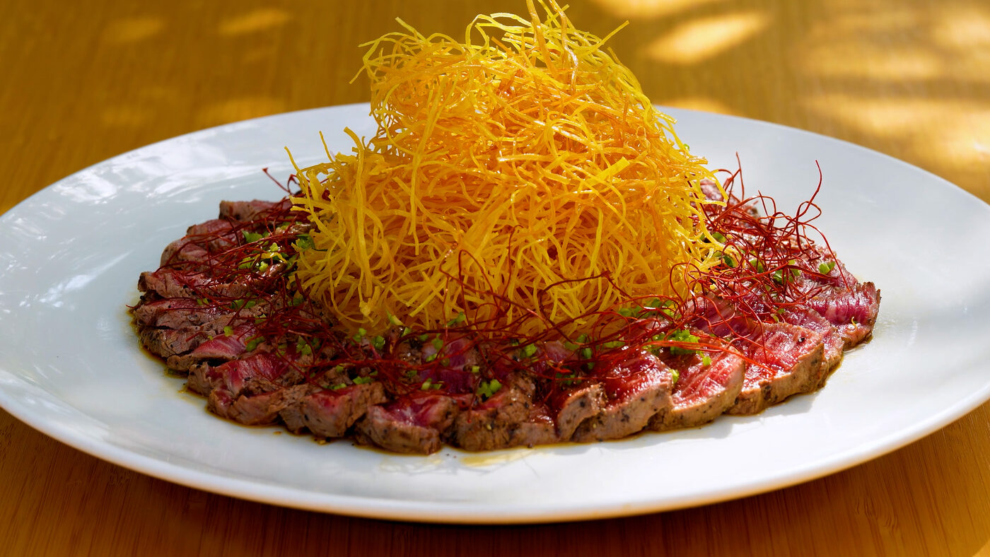 Sliced seared beef tartare topped with crispy golden potato strings on a white plate