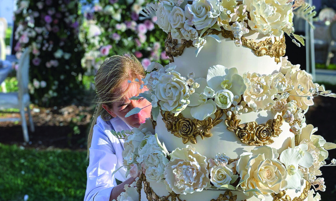 Julie Deffense adding finishing touches to a tall, ornate white and gold wedding cake outdoors